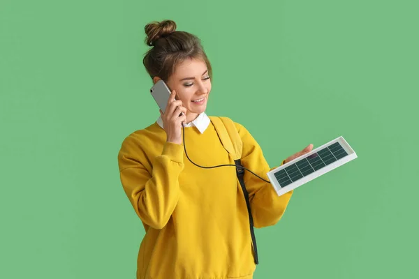 Pretty young woman charging mobile phone with portable solar panel on green background