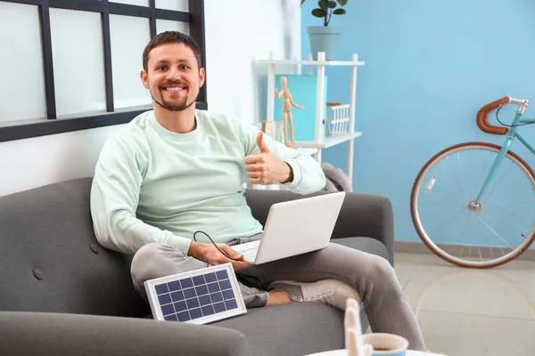 Handsome man with laptop charging from portable solar panel showing thumb-up at home