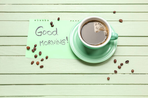 Cup of coffee, beans and paper with text GOOD MORNING on color wooden background