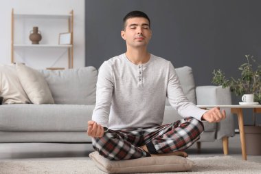 Young man in pajamas meditating on pillow at home