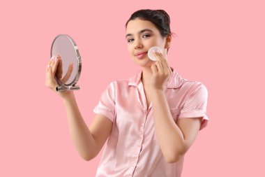 Aynalı genç bir kadın pembe arka planda makyajı kaldırıyor.