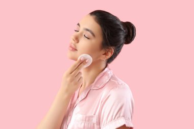 Genç bir kadın pembe arka planda pamuk yastıkla makyajı kaldırıyor.