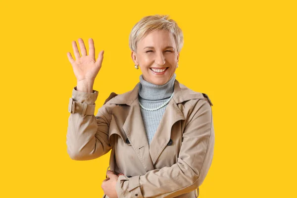 Beautiful Mature Woman Coat Waving Hand Yellow Background — Stock Photo, Image