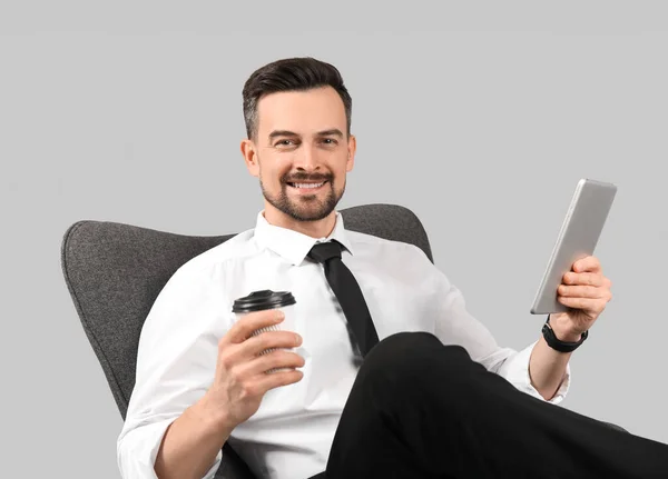 灰色の背景にアームチェアにタブレットコンピュータとコーヒーカップを持つハンサムなビジネスマン — ストック写真
