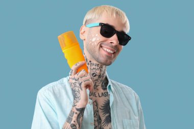 Açık mavi arka planda güneş kremi sürmüş dövmeli bir adam.