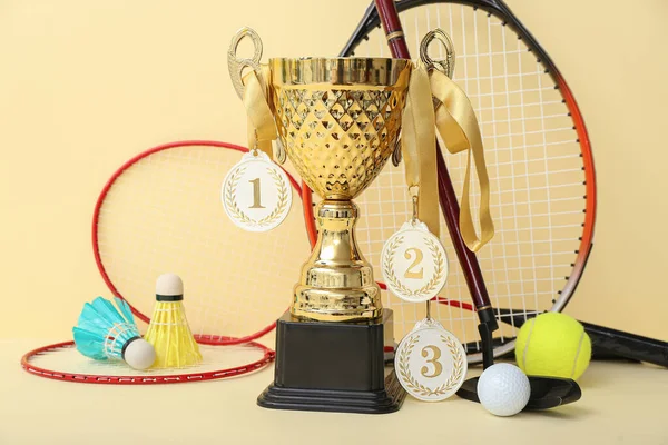 Gold cup with prize medals and sports equipment on yellow background