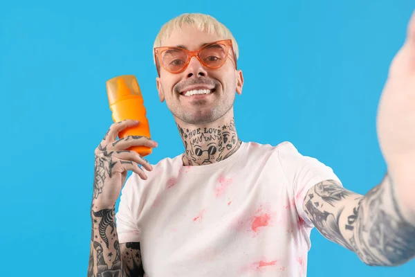 Tattooed man with sunscreen cream taking selfie on light blue background