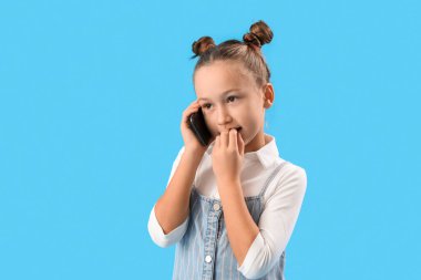 Little girl biting nails while talking by mobile phone on blue background