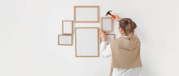 stock image Young woman with hammer hanging blank photo frames on light wall