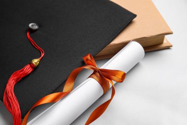 Kırmızı kurdeleli diploma, mezuniyet şapkası ve beyaz arka plan kitabı.