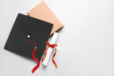 Kırmızı kurdeleli diploma, mezuniyet şapkası ve beyaz arka plan kitabı.