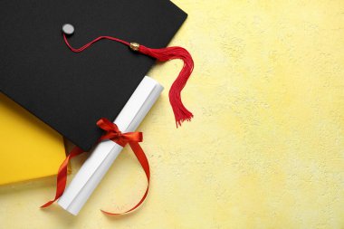 Kırmızı kurdeleli diploma, mezuniyet şapkası ve sarı masadaki kitap.