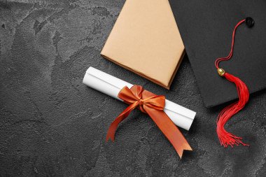 Kırmızı kurdeleli diploma, mezuniyet şapkası ve siyah masa üzerine kitap.
