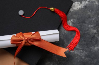 Kırmızı kurdeleli diploma, mezuniyet şapkası ve koyu gri masa üzerine kitap.