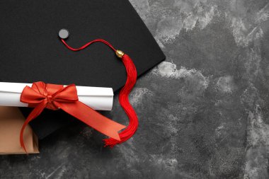 Kırmızı kurdeleli diploma, mezuniyet şapkası ve koyu gri masa üzerine kitap.