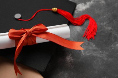 Kırmızı kurdeleli diploma, mezuniyet şapkası ve koyu gri masa üzerine kitap.