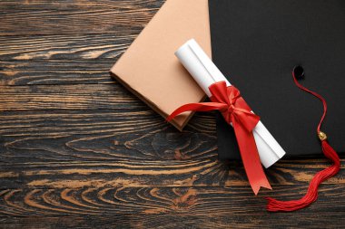 Kırmızı kurdeleli diploma, mezuniyet şapkası ve ahşap masa üzerine kitap.
