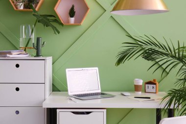 Ofiste yeşil duvarın yanında dizüstü bilgisayarı ve kahve fincanı olan bir iş yeri.