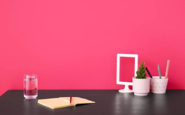 Kırmızı duvarın yanında not defteri, kalem bardağı ve bir bardak su olan işyeri