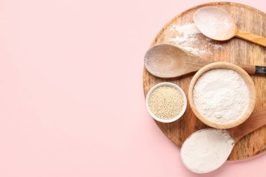 Üzerinde buğday unu, susam tohumu ve pembe arka planda kaşıklar olan bir tahta.