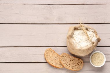 Çuval dolusu buğday unu, susam tohumları ve gri ahşap masada bir parça ekmek.