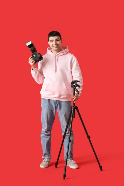 赤の背景にプロのカメラと三脚を持つ若い男性写真家 — ストック写真