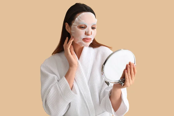 Young woman with sheet mask looking in mirror on beige background