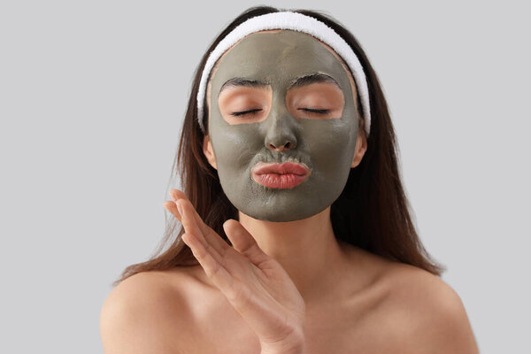 Young woman with applied clay mask blowing kiss on grey background, closeup