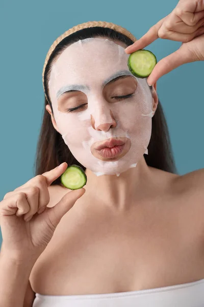 Young woman with sheet mask and cucumber slices blowing kiss on blue background, closeup