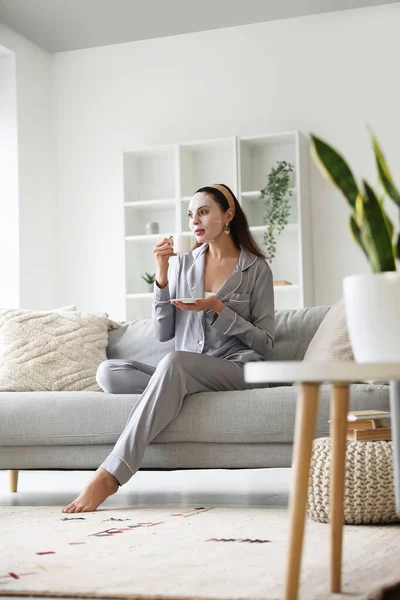 Young woman with sheet mask and cup of coffee sitting on sofa at home
