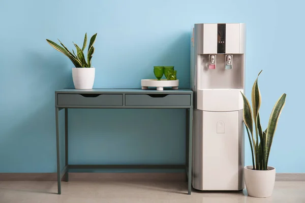 Modern water cooler, table, glasses and houseplants near blue wall