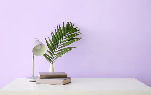 Workplace with lamp, books and palm leaf near lilac wall