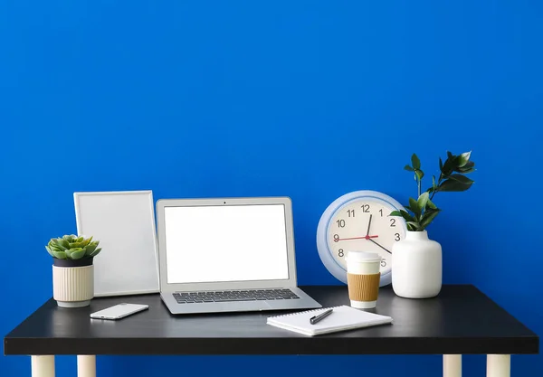 Workplace with laptop, frame, cup of coffee and clock near blue wall