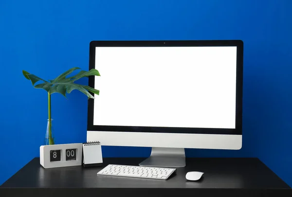 Workplace with computer, clock and palm leaf near blue wall