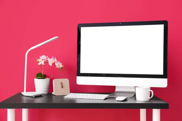 Workplace with computer, clock, flower and lamp near red wall
