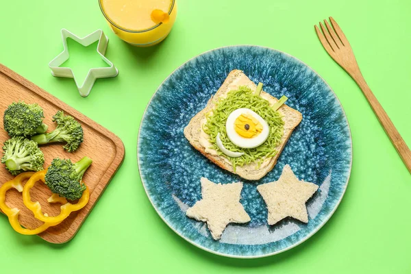 Plate with funny breakfast in shape of alien, vegetables and glass of juice on green background