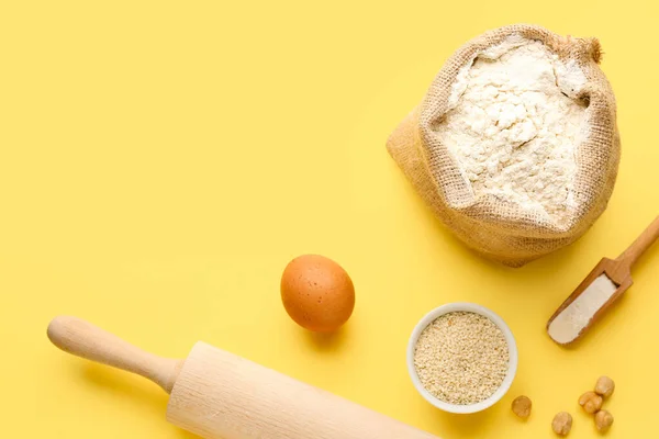 Composition with sack bag of wheat flour, sesame seeds, egg and rolling pin on yellow background