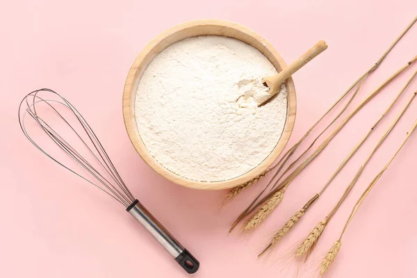 Bowl with flour, wheat ears and whisk on pink background