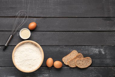 Buğday unu, yumurta, ekmek parçaları ve siyah ahşap masa üzerinde çırpma kabı.