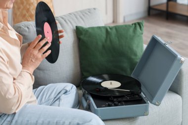 Evde kanepede oturan, plakçalar için vinil diski olan olgun bir kadın.