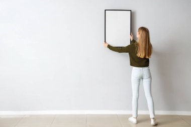 Young woman hanging blank frame on light wall at home, back view