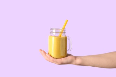 Mason kavanozunda pembe arka planda sarı smoothie olan bir kadın eli.