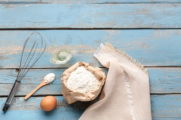 Bag of wheat flour, egg, oil, whisk and spoon on blue wooden table