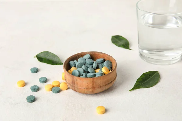 stock image Bowl with pills, leaves and glass of water on white table