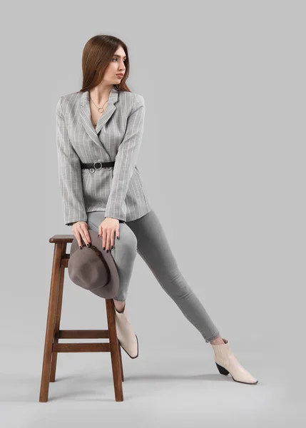 穿着夹克和牛仔裤的时髦年轻女子坐在灰色背景的凳子上 — 图库照片
