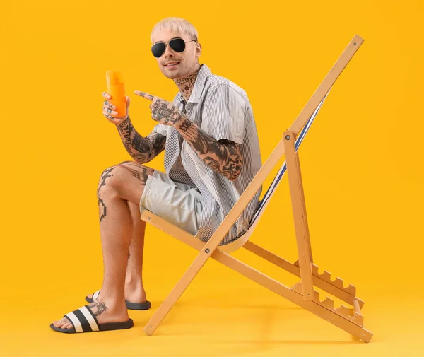 Tattooed man with sunscreen cream sitting on deck chair against yellow background