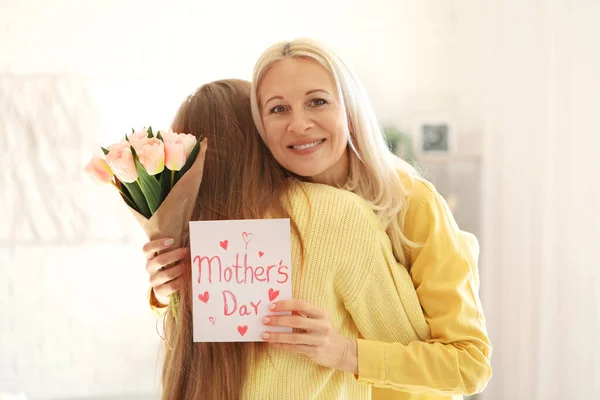 Mature woman with tulips and card for Mother\'s Day hugging her daughter at home