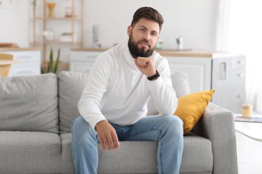 Thoughtful young bearded man sitting on sofa in kitchen
