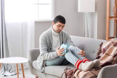 Young man warming his knee with hot water bottle on sofa at home