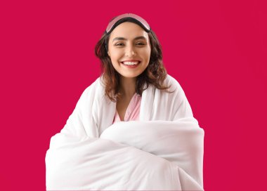 Young brunette woman with sleeping mask and blanket on pink background